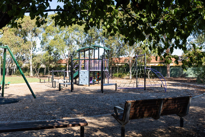 Shaded bench seats looking towards a children's playground with climbing structures and swings.