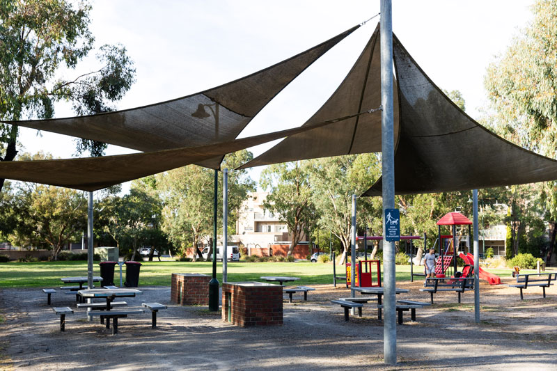 4 picnic tables and two brick BBQs sheltered by 4 overlapping shade sails, beside a playground with benches.