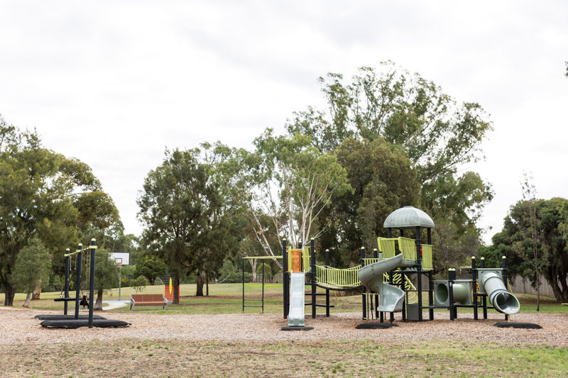 Children's playground with climbing structures, slides and swings with large trees in the background.