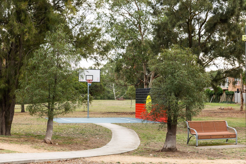 A concrete path leads to the half basketball court with Australian Aboriginal Flag mural on the rebounding wall.