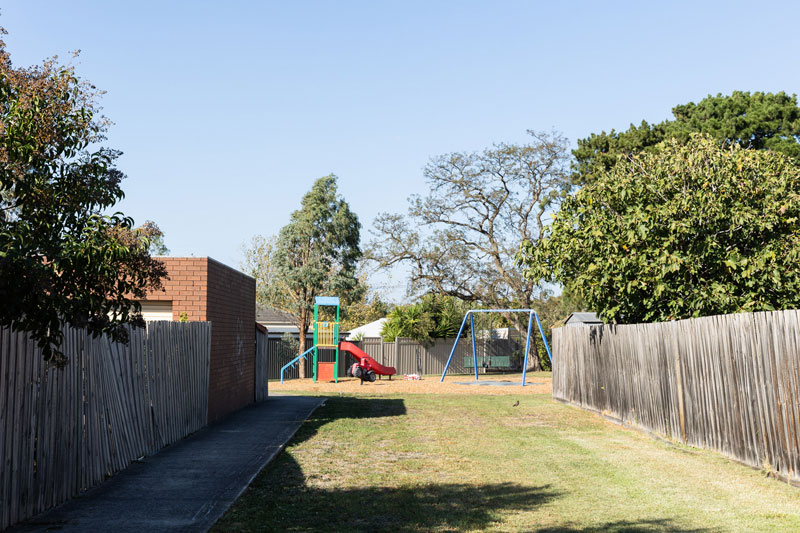 A small playground can be seen at the end of a grassy walkway between two suburban, side fences.