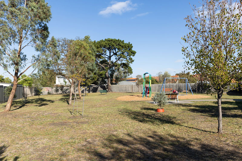 A small park is surrounded by suburban wooden fences, with green open space, scattered trees and a small playground.