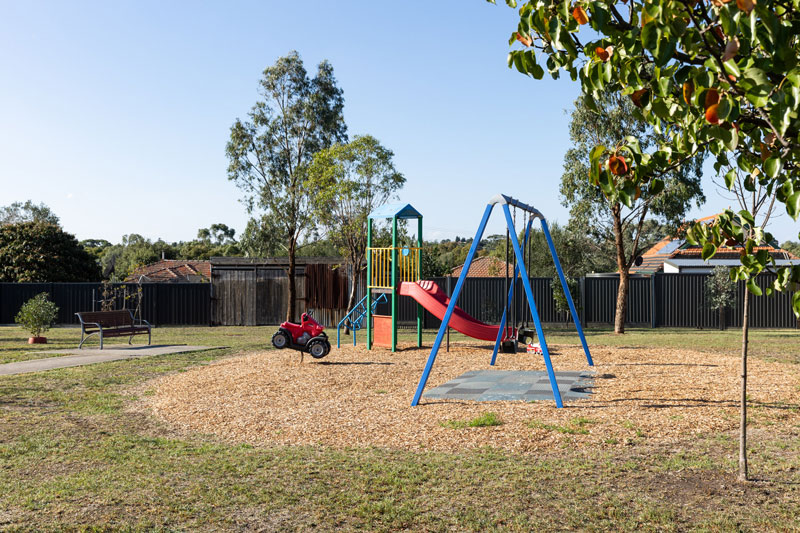 A park bench faces a small colourful playground with a slide, swing set and a racker in the shape of a red car.