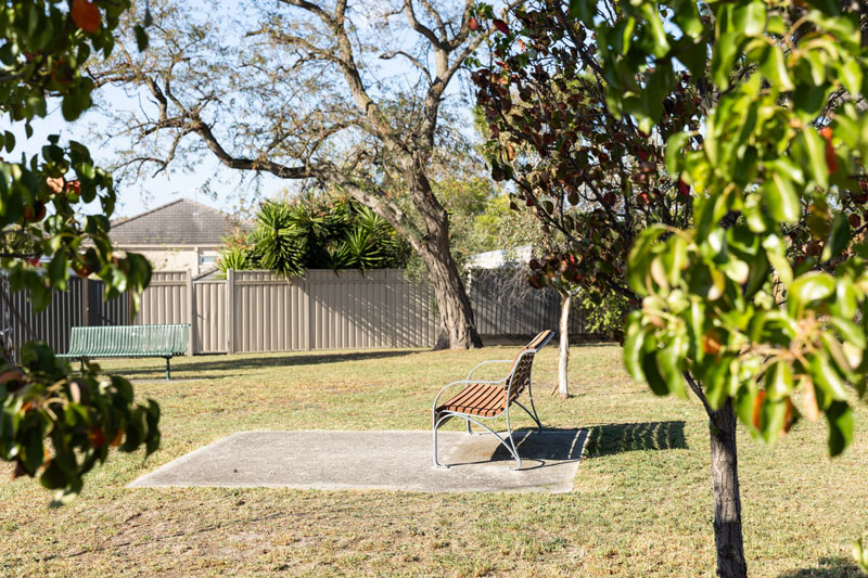 Two park benches can be seen in a pocket park, one in green metal and the other with a wooden seat.
