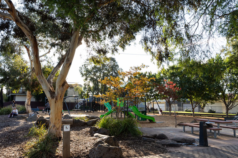 Families enjoy the park where a bright green playground can be seen through the trees and there are picnic tables and a drinking tap in the foreground.