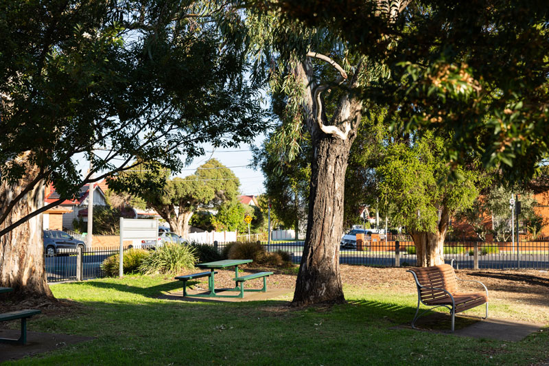 The corner of a fenced park in the afternoon light has 2 green picnic tables and a bench seat under mature trees.