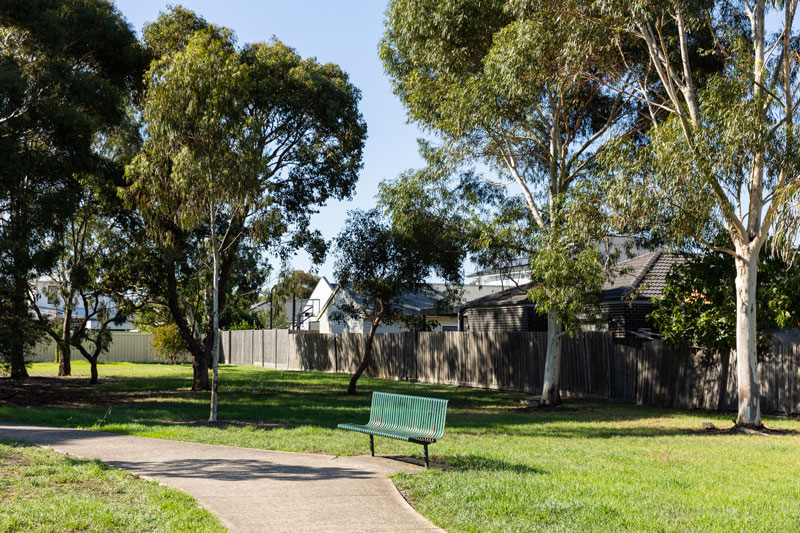 A park bench seat next to the concrete pathway, next to a lawn shaded by tall native trees.