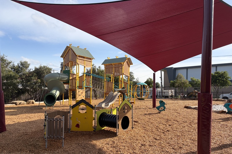 Playground structures for young children underneath red sail shade.