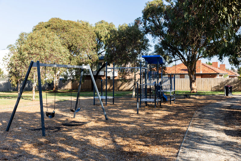 A children's playground with swings and climbing structures shaded by large native trees.