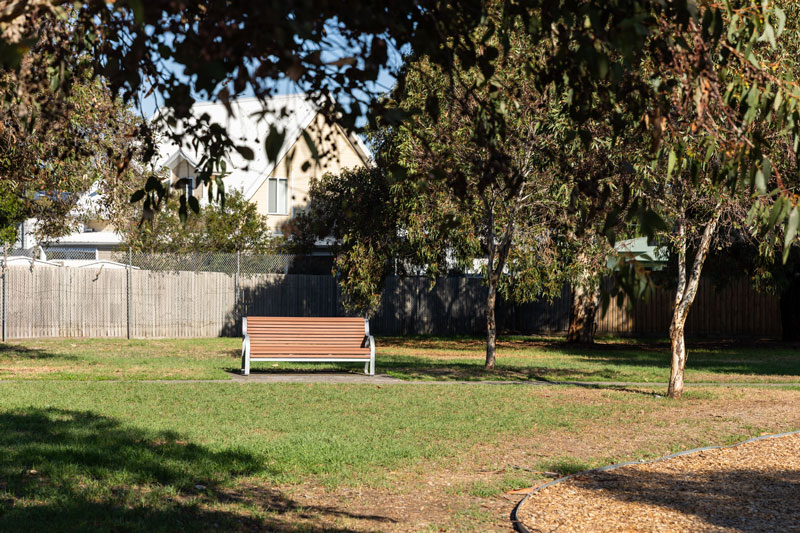 A park bench seat next to a concrete pathway in lawned area with native trees.